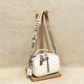 Cartera de silueta tipo "camera bag" con textura acolchada en patrón de diamantes. Incluye manilla superior rígida, cierres metálicos con dijes decorativos y una correa ancha de tela ajustable con la palabra "LOVE" grabada.