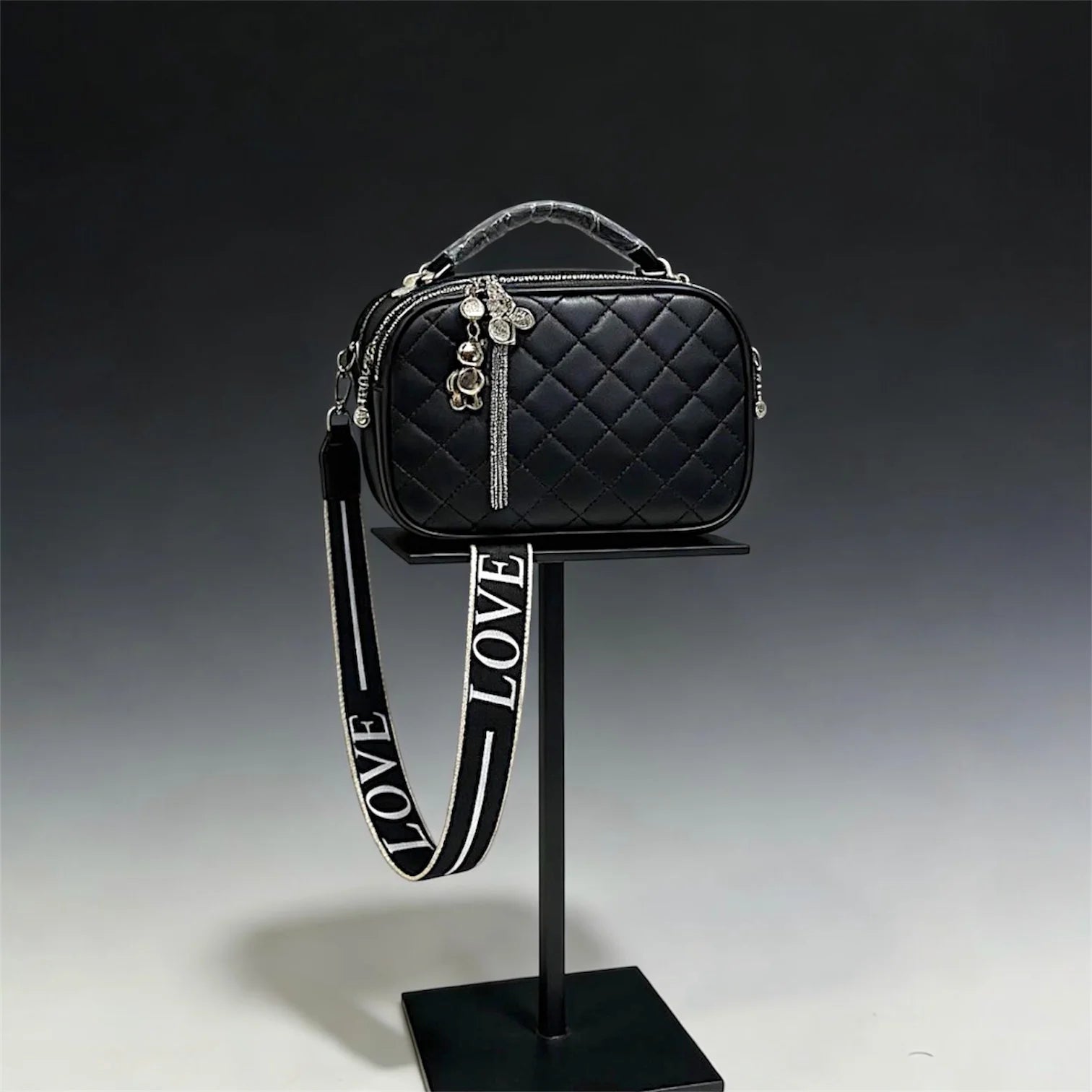 Cartera de silueta tipo "camera bag" con textura acolchada en patrón de diamantes. Incluye manilla superior rígida, cierres metálicos con dijes decorativos y una correa ancha de tela ajustable con la palabra "LOVE" grabada.