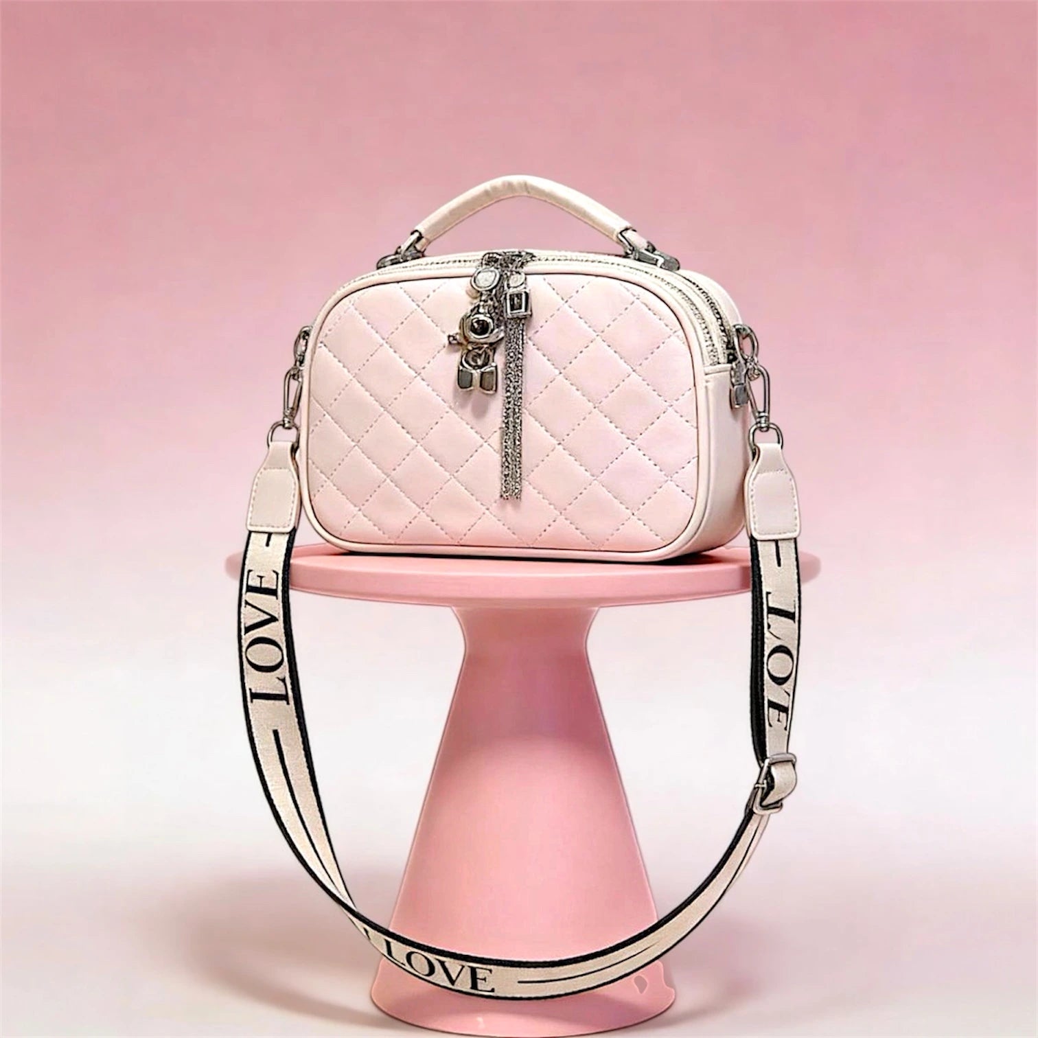 Cartera de silueta tipo "camera bag" con textura acolchada en patrón de diamantes. Incluye manilla superior rígida, cierres metálicos con dijes decorativos y una correa ancha de tela ajustable con la palabra "LOVE" grabada.