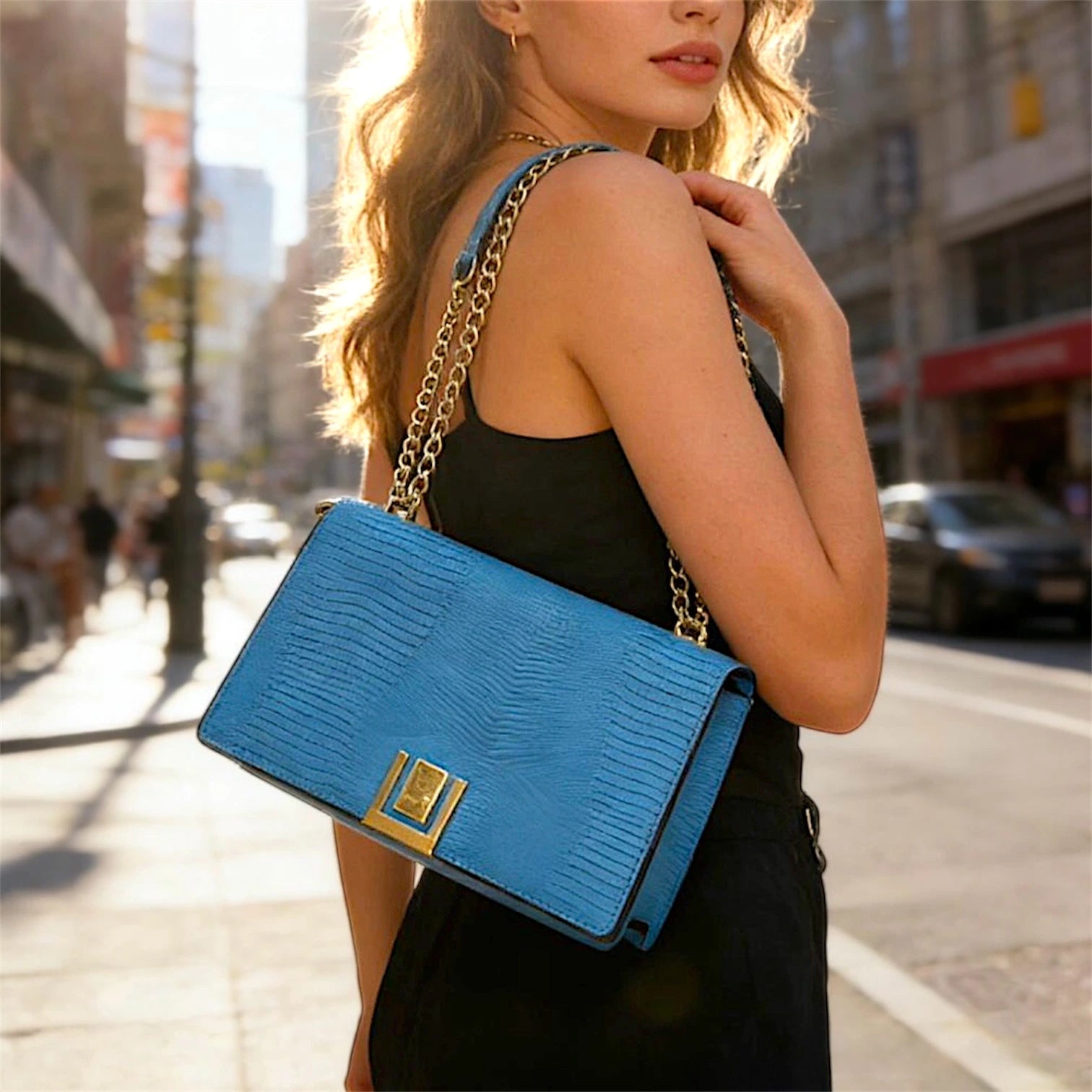 Cartera de hombro estructurada con solapa frontal en textura de piel de lagarto (lizard). Presenta un cierre metálico dorado rectangular, correa de cadena dorada entrelazada y un diseño compacto de líneas limpias.