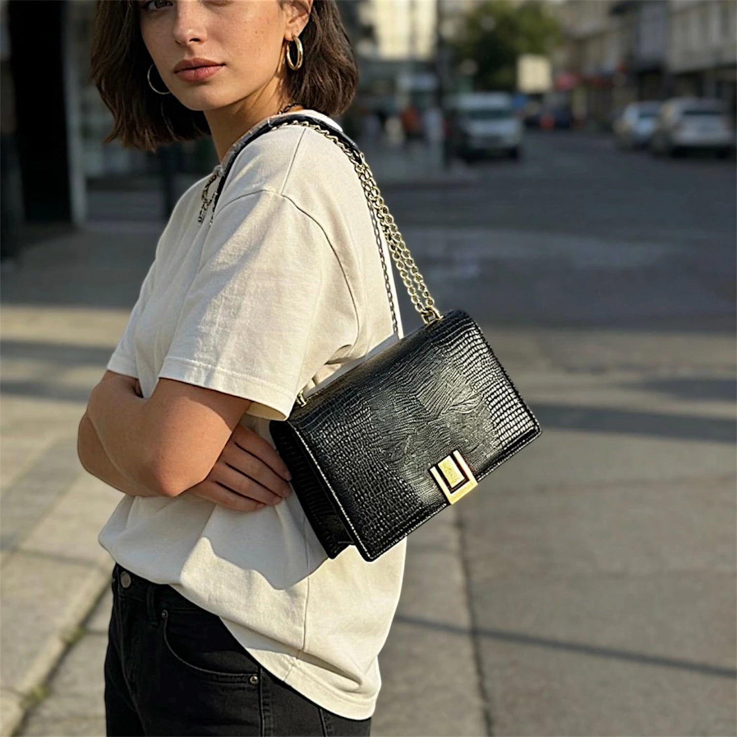 Cartera de hombro estructurada con solapa frontal en textura de piel de lagarto (lizard). Presenta un cierre metálico dorado rectangular, correa de cadena dorada entrelazada y un diseño compacto de líneas limpias.