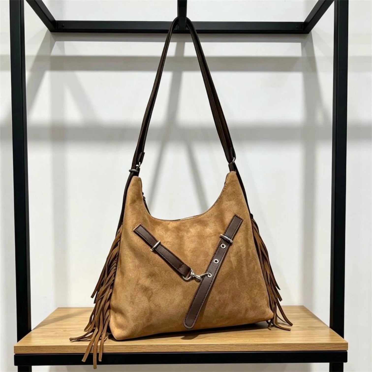 Cartera negra de gamuza con flecos laterales y correa cruzada con hebillas plateadas, colgada en un exhibidor metálico.