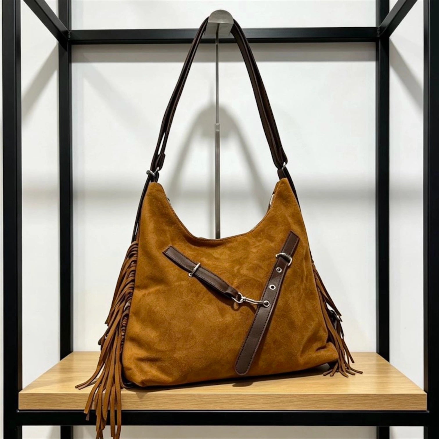 Cartera negra de gamuza con flecos laterales y correa cruzada con hebillas plateadas, colgada en un exhibidor metálico.