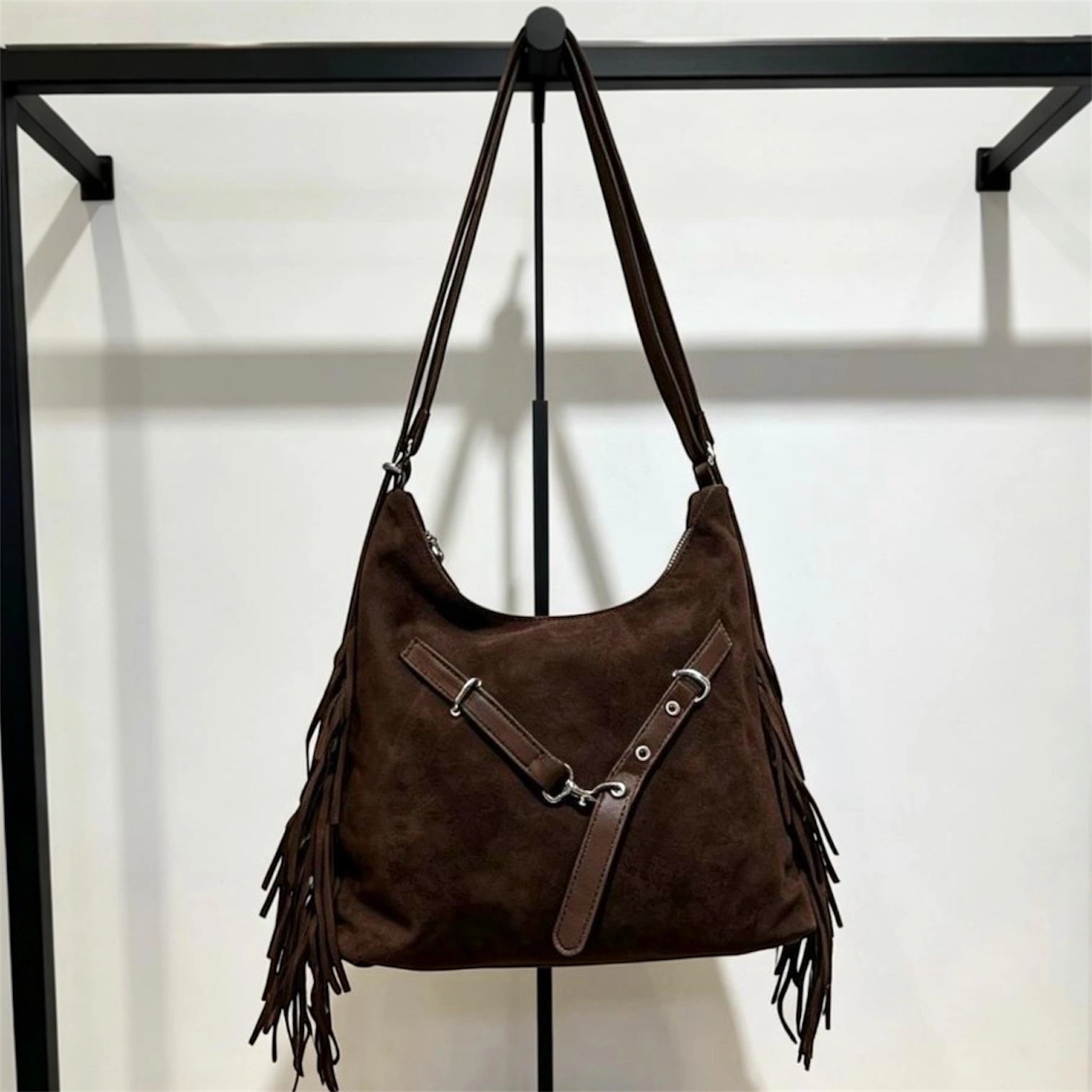 Cartera negra de gamuza con flecos laterales y correa cruzada con hebillas plateadas, colgada en un exhibidor metálico.