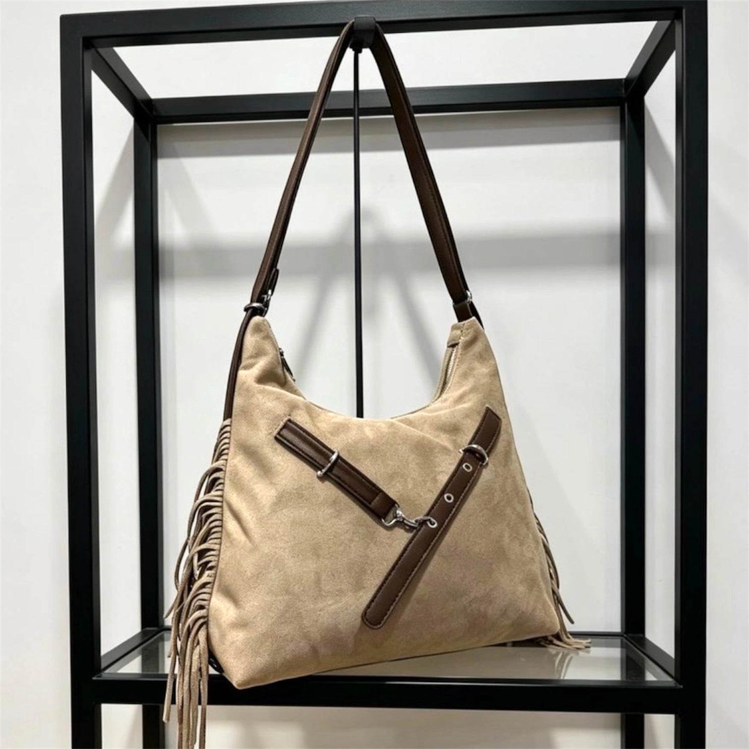 Cartera negra de gamuza con flecos laterales y correa cruzada con hebillas plateadas, colgada en un exhibidor metálico.